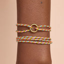 Multicolored braided bracelet with a gold ring on a wrist against a beige background